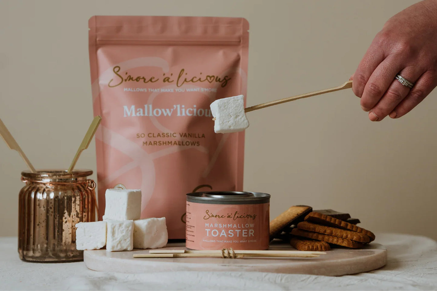 Mallowlicious marshmallow products on a table with a hand toasting a marshmallow.